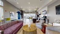 Located at 9050 W Chester Blvd in West Chester, Ohio, this single-floor community clubhouse offers a generously sized, thoughtfully designed gathering space that blends modern aesthetics with functional versatility. The interior is anchored by a soaring reception lounge featuring dramatic pendant lighting, light wood-look flooring, bold abstract artwork, and deep blue velvet accent chairs arranged around a marble-top table — creating a welcoming first impression.

The open-concept social lounge transitions seamlessly into a café-style amenity area equipped with a full-length bar counter, refrigerator, coffee station, and multiple wall-mounted televisions. A striking industrial-style overhead garage door opens fully to a covered outdoor terrace furnished with plush blue seating, a ceiling fan, and views of the surrounding community — effortlessly blending indoor and outdoor living.

A dedicated lounge zone features vibrant yellow-green bar stools, high-top tables, and cozy booth-style seating beside tall windows that flood the space with natural light. A private conference room, carpeted and outfitted with a long wooden table and plum-toned executive chairs, provides a quiet setting for meetings. Throughout, warm wood accent walls, recessed lighting, and a curated color palette of gray, magenta, and gold create a cohesive, upscale ambiance.