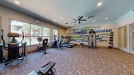 This fitness center offers a comprehensive workout environment designed to support a wide range of exercise routines. The space is anchored by a vibrant, eye-catching word-art mural spanning an entire wall, featuring fitness-themed typography in bold blue and green tones that energizes the atmosphere throughout. Recessed lighting and a ceiling fan keep the space well-lit and comfortable during workouts.

The cardio section is positioned near large windows that overlook the surrounding outdoor area, allowing natural light to fill the space while providing an pleasant view. Equipment includes treadmills, elliptical trainers, and stationary bikes to accommodate various cardio preferences.

The strength training area is outfitted with a full dumbbell rack, a multi-station cable machine, a Smith machine with weight plates, a weight bench, and additional resistance machines for targeted muscle training. Floor-to-ceiling mirrors line one wall, enhancing the sense of openness and allowing users to monitor their form. Medicine balls and a kettle bell round out the free weight offerings.

Additional amenities include wall-mounted water fountains for convenient hydration and a clearly posted fitness rules board near the entrance. The durable, patterned carpet flooring ties the space together, creating a functional and motivating environment well-suited for daily use.