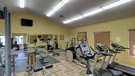 This single-level fitness room offers a comprehensive workout environment designed to accommodate a variety of exercise routines. The space is finished with warm yellow walls that create an energizing atmosphere, complemented by a parquet-style flooring that adds a classic touch throughout. Overhead fluorescent lighting ensures the room is well-lit at all times.

The cardio section is prominently featured, housing multiple treadmills, an elliptical trainer, and a stationary bike, all positioned to allow comfortable use and easy navigation between machines. A dedicated strength and stretching area includes a weight bench with a padded mat, free weights, and additional resistance equipment. Large wall mirrors along one side of the room enhance the sense of space while allowing users to monitor their form during workouts.

A wall-mounted television provides entertainment for those engaged in longer cardio sessions. A window fitted with wooden blinds allows for ventilation and a connection to the outside. The overall layout is practical and organized, making efficient use of the available floor space to support a well-rounded fitness routine.