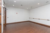 Located at 2071 Powers Ferry Rd SE in Marietta, Georgia, this professional dance studio offers a well-appointed, single-floor space designed to support a variety of dance disciplines. The studio's most prominent features are its wall-mounted wooden ballet barres, which extend along multiple walls, providing practitioners ample room for warm-ups and technique work. Floor-to-ceiling mirrors line one side of the studio, creating a sense of expanded space while allowing dancers to monitor and refine their form. The open floor plan is finished with rich, dark hardwood flooring that adds warmth and a classic studio aesthetic, while also providing the smooth, resilient surface essential for dance practice. Recessed ceiling lighting illuminates the space evenly, ensuring clear visibility throughout. Crisp white walls and clean dark wood baseboards complement the flooring and contribute to a polished, professional atmosphere. Elegant dark wood double doors serve as the main entry point, lending a refined character to the space and flanked by additional single doors for convenient access. The layout is open and unobstructed, maximizing usable floor space for classes, rehearsals, or individual practice. Overall, this studio combines functional design with a clean, professional ambiance well-suited for dance instruction and performance preparation.