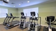 This fitness center offers a dedicated, single-floor space designed to support a well-rounded cardio workout routine. The room is finished with soft green walls accented by crisp white crown molding and clean white baseboards, creating a calm and motivating environment. Recessed lighting in the ceiling provides consistent, even illumination throughout the space, while a large window fitted with white blinds allows natural light to fill the room, enhancing the open feel of the interior. The flooring features a combination of low-pile carpet and protective rubber mats beneath the equipment, offering both comfort and durability. The fitness area is outfitted with professional-grade TRUE brand elliptical cross-trainers and treadmills, providing users with high-quality options for cardiovascular exercise. The machines are thoughtfully arranged to allow ample space between each unit, ensuring a comfortable and safe workout experience. The overall design of the space is practical and well-maintained, with a clean aesthetic that prioritizes functionality. Whether used for a light walk or an intense training session, this fitness center provides the equipment and environment needed to support a consistent and effective exercise routine.
