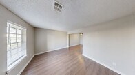 This two-bedroom, one-bathroom two-story home offers a well-organized layout spread across two floors. The main floor houses the combined kitchen and living area, a laundry room, and a connecting hallway. The open living room features warm hardwood-style flooring, a ceiling fan with light fixture, and a large window that fills the space with natural light. A half-wall pass-through connects the living area to the kitchen, which is equipped with white shaker-style cabinetry with black hardware, a double-basin sink with a matte black faucet, white countertops, an under-cabinet range hood, and a skylight-style overhead light fixture. The laundry room is conveniently accessible directly from the kitchen. The upper floor accommodates both bedrooms and the full bathroom. Each bedroom features hardwood-style flooring, neutral wall tones, ceiling fans, and windows with white blinds that invite natural light. The primary bedroom is generously sized with a closet and a window overlooking the exterior. The shared bathroom includes a tub-shower combo with clean white subway tile surround and glass corner shelving. Textured ceilings are present throughout, adding a classic character to the home's overall clean and neutral aesthetic.