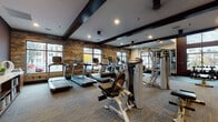 This fitness center offers a comprehensive and thoughtfully designed workout environment spread across a single level. The space is defined by a striking combination of rustic reclaimed wood accent walls and dark exposed ceiling beams, creating a modern-industrial aesthetic that sets it apart from a conventional gym setting. Patterned carpet flooring with yellow accent lines spans the main workout area, while rubber matting is placed beneath heavy equipment for added stability.

The cardio zone lines one side of the space, housing multiple Matrix treadmills equipped with individual digital screens, an elliptical trainer, and a row of spin bikes positioned near large windows that overlook the surrounding streetscape, allowing natural light to fill the area during workouts. A dedicated free weight section features a tiered dumbbell rack positioned beneath a floor-to-ceiling window, offering ample natural illumination. Mirrored walls expand the visual depth of the strength training area, which includes a cable machine system and multi-use functional trainer. Additional equipment such as stability balls and foam rollers are neatly stored on built-in wooden shelving. Recessed lighting throughout ensures consistent brightness, complementing the natural light provided by the numerous large-pane windows.