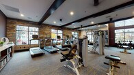 This fitness center offers a comprehensive and thoughtfully designed workout environment spread across a single level. The space is defined by a striking combination of rustic reclaimed wood accent walls and dark exposed ceiling beams, creating a modern-industrial aesthetic that sets it apart from a conventional gym setting. Patterned carpet flooring with yellow accent lines spans the main workout area, while rubber matting is placed beneath heavy equipment for added stability.

The cardio zone lines one side of the space, housing multiple Matrix treadmills equipped with individual digital screens, an elliptical trainer, and a row of spin bikes positioned near large windows that overlook the surrounding streetscape, allowing natural light to fill the area during workouts. A dedicated free weight section features a tiered dumbbell rack positioned beneath a floor-to-ceiling window, offering ample natural illumination. Mirrored walls expand the visual depth of the strength training area, which includes a cable machine system and multi-use functional trainer. Additional equipment such as stability balls and foam rollers are neatly stored on built-in wooden shelving. Recessed lighting throughout ensures consistent brightness, complementing the natural light provided by the numerous large-pane windows.