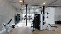 This single-floor fitness space showcases a refined industrial aesthetic, combining raw architectural elements with a clean, functional layout. The exposed concrete ceiling features visible ductwork and structural elements, lending the space an authentic, loft-like character. Black steel support columns punctuate the open floor plan, reinforcing the industrial design language while providing structural definition throughout the workout area.

Natural light floods the interior through large windows at the far end of the space, complemented by a series of skylights embedded in the ceiling. This combination creates a bright, energizing atmosphere well-suited for training. Warm-toned hardwood flooring provides a striking contrast against the monochromatic palette of the ceiling and equipment, adding visual warmth to the otherwise utilitarian environment.

The space is equipped with professional-grade strength machines, including a cable and pulley system and a dedicated resistance training unit. Full-length mirrors line one wall, a practical feature that supports proper form during workouts while also amplifying the sense of openness. The overall layout is spacious and well-organized, allowing for comfortable movement between stations.