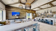 Located at 16100 Myriad Ln in Fort Myers, FL, this lobby and clubhouse is a thoughtfully designed community gathering space that blends coastal aesthetics with modern comfort. The single-floor layout spans a generous footprint, encompassing multiple distinct areas that flow seamlessly into one another.

The main lounge area is anchored by soaring vaulted ceilings with dark exposed beams and oversized woven pendant light fixtures that add a warm, organic texture. A dramatic floor-to-ceiling wood-panel accent wall serves as a focal point, complemented by a large wall-mounted display. Ample natural light pours in through expansive sliding glass doors and large windows, framing views of the outdoor pool and surrounding tropical landscaping.

The space transitions into a casual seating and dining area featuring a long communal table with stylish upholstered chairs, pendant lighting, and decorative coastal artwork. A kitchenette counter with bar seating provides additional functionality for social gatherings.

The entry lobby greets visitors with tall indoor palms, elegant crystal chandelier fixtures, patterned bench seating, and glass-partitioned interiors that lend an open, airy feel. Throughout, large-format light tile flooring ties each zone together, enhancing the bright and inviting atmosphere that defines this well-appointed Fort Myers clubhouse.