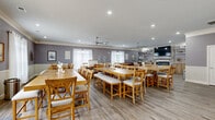 Located at 445 Warfield Blvd in Clarksville, Tennessee, this single-floor community space offers a generously sized, open-concept layout designed to accommodate group dining, relaxation, and social gatherings. The interior is finished with warm, neutral tones — soft gray walls, white wainscoting, and crown molding — complemented by light-toned luxury vinyl plank flooring that flows consistently throughout.

The expansive dining area is the centerpiece of the space, featuring multiple long farmhouse-style wooden tables with cross-back chairs and upholstered benches, all illuminated by recessed lighting and ceiling fans. Sheer white curtains on large windows invite ample natural light, creating a bright and welcoming atmosphere.

A full kitchen anchors one end of the space, outfitted with light wood cabinetry, a subway tile backsplash, stainless steel appliances, and a striking quartz-topped island with pendant lighting. Open shelving adds both function and style.

Adjacent to the kitchen is a cozy lounge area featuring a large sectional leather sofa, a wall-mounted TV, a brick-surround fireplace, and a built-in beverage and storage station with wood cabinetry. A more intimate dining nook with a single table and chairs provides a quieter setting. Together, these zones create a versatile, community-focused environment ideal for shared living or event programming.