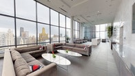 This upscale high-rise lounge space delivers a striking first impression through its expansive floor-to-ceiling curtain-wall glazing, which frames breathtaking panoramic views of the Chicago skyline — including iconic landmarks such as the Willis Tower and Tribune Tower. The open-plan layout is generously proportioned, with large-format porcelain tile flooring in soft grey tones running throughout, lending the space a cohesive, polished character.

The main lounge area is furnished with plush, contemporary sectional sofas and modular lounge chairs in neutral taupe upholstery, arranged around sleek oval white coffee tables. Multiple seating zones throughout the floor encourage both social gathering and quiet retreat, each positioned to maximize the dramatic cityscape backdrop. Slender cylindrical columns punctuate the space without interrupting sightlines.

A dedicated reception area anchors one end of the floor, featuring clean-lined millwork cabinetry in warm wood tones set against a soaring wall clad in veined marble-effect stone. Wall-mounted flat screens and pendant lighting complete the professional yet welcoming environment.

Natural light floods the interior throughout the day, reflecting off the light-toned surfaces and reinforcing the airy, open ambiance. The overall aesthetic is one of refined urban sophistication — a seamless blend of functionality and contemporary design at an elevated vantage point above the city.