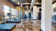 This fitness center combines industrial aesthetics with a fully functional workout environment. The space is defined by its exposed brick walls, which line the perimeter and frame a row of large windows that flood the interior with natural light and offer outdoor views of landscaped surroundings. Polished concrete floors with a high-gloss finish run throughout, complementing the dark ceiling accents and exposed silver ductwork that give the space an urban, warehouse-inspired character.

The cardio zone features multiple treadmills positioned directly in front of the large windows, providing an energizing view during workouts. A dedicated strength and conditioning area includes adjustable benches, a dumbbell rack, and rowing machines, with full-length mirrors along the walls to support proper form. Motivational typography — including phrases like "Grit. Power. Strength." and "Be Stronger Than Your Excuses" — is displayed prominently on the walls, reinforcing the space's athletic atmosphere.

Additional amenities include a bank of lockers for secure storage, a wall-mounted television, and a water station. Recessed lighting supplements the abundant natural light, ensuring the space remains well-lit throughout the day. The open floor plan allows for flexible use across multiple fitness disciplines.