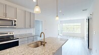 This two-bedroom, two-bathroom residence offers a thoughtfully designed layout spread across two floors, combining modern finishes with a bright, open aesthetic. The kitchen serves as a central highlight, featuring granite countertops, a large center island with a stainless steel sink and brushed gold faucet, gray shaker-style cabinetry extending to the ceiling, and a full suite of stainless steel appliances. Pendant lighting adds a refined touch above the island. The kitchen flows seamlessly into the living room, which is bathed in natural light from expansive windows overlooking lush greenery, with light hardwood-style flooring tying the spaces together. A private balcony accessible from the living area offers serene views of a manicured lawn, mature trees, and a wooden fence line. The primary bedroom is generously sized, with soft carpet flooring, a large window framing outdoor views, and direct access to a spacious walk-in closet equipped with wire shelving. The second bedroom is equally comfortable, also featuring carpet and ample natural light. Both bathrooms are clean and functional, with one including a full tub-shower combination fitted with brushed gold fixtures. A hallway and dedicated closet complete the floor plan, providing practical storage and circulation throughout the home.