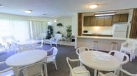 This single-floor community lounge is designed to serve as a welcoming gathering space for residents and guests alike. The room is bathed in natural light streaming through large floor-to-ceiling windows dressed with vertical blinds, creating an open and airy atmosphere throughout.

The layout is thoughtfully organized into distinct seating areas. Multiple round white tables paired with white plastic chairs are arranged to accommodate group dining or casual conversation. A separate lounge area features elegant white wicker chairs and a loveseat accented with blue cushions, offering a more relaxed setting. Lush potted plants are strategically placed throughout the room, adding warmth and a touch of nature to the interior.

A ceiling fan helps maintain comfortable airflow, complementing the overall light and pleasant ambiance. The space features a textured ceiling and plush carpeted flooring in a muted blue-gray tone that ties the room together cohesively.

Along one wall, a built-in kitchenette provides added functionality, complete with wood-toned upper cabinetry, a countertop pass-through, a microwave, and a sink — making it convenient for hosting events or preparing light refreshments. A bar-height stool at the counter adds a casual dining option. The overall design is clean, practical, and inviting, well-suited for community activities and social interaction.