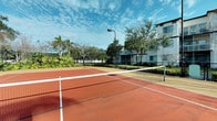 This outdoor recreational facility centers around a full-sized tennis court finished in a rich terracotta clay-style surface, complete with crisp white boundary lines and a regulation net. The court is fully enclosed by chain-link fencing, ensuring safety and defining the play area while maintaining an open, airy feel. Overhead court lighting is provided by a tall mounted lamp post, allowing for extended play beyond daylight hours. The surrounding landscape is verdant and well-kept, with a variety of mature tropical trees and dense shrubbery that create a natural, park-like atmosphere. The lush greenery provides both shade and a visually appealing backdrop that enhances the overall ambiance of the space. A modern multi-story residential building is visible just beyond the court's perimeter, indicating that this facility serves as a community amenity. The open sky above allows for ample natural light throughout the day, complemented by the airy tropical setting. The combination of a quality playing surface, thoughtful lighting, and beautifully maintained landscaping makes this tennis court a standout recreational feature within the community.