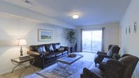 This single-floor space centers around a thoughtfully designed living room that combines comfort and style. The room is furnished with dark leather seating, including a sofa and accent chairs, arranged around a sleek glass-top coffee table resting on a patterned area rug. A framed landscape artwork adorns the wall, adding a decorative focal point to the space. A tall indoor plant positioned near the sliding glass door introduces a natural element to the interior. The living room benefits from ample natural light streaming through large sliding glass doors dressed with draping curtains, which open directly onto an outdoor patio area, seamlessly connecting indoor and outdoor living. The neutral-toned carpeted flooring and light-colored walls contribute to an airy, open ambiance throughout. Overhead, a flush-mount ceiling light provides consistent illumination, complementing the natural light. Crown molding along the ceiling adds a refined architectural detail to the room. Overall, the space presents a clean, welcoming atmosphere with functional furnishings and well-considered design elements.