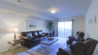 This single-floor space centers around a thoughtfully designed living room that combines comfort and style. The room is furnished with dark leather seating, including a sofa and accent chairs, arranged around a sleek glass-top coffee table resting on a patterned area rug. A framed landscape artwork adorns the wall, adding a decorative focal point to the space. A tall indoor plant positioned near the sliding glass door introduces a natural element to the interior. The living room benefits from ample natural light streaming through large sliding glass doors dressed with draping curtains, which open directly onto an outdoor patio area, seamlessly connecting indoor and outdoor living. The neutral-toned carpeted flooring and light-colored walls contribute to an airy, open ambiance throughout. Overhead, a flush-mount ceiling light provides consistent illumination, complementing the natural light. Crown molding along the ceiling adds a refined architectural detail to the room. Overall, the space presents a clean, welcoming atmosphere with functional furnishings and well-considered design elements.