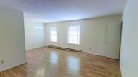 This single-floor space offers a clean and open layout that is both inviting and functional. The main living area is a standout feature, boasting gleaming light-toned hardwood floors that reflect natural light beautifully throughout the room. Two generously sized windows dressed with white horizontal blinds allow ample daylight to fill the space, creating a bright and airy atmosphere. A classic multi-arm chandelier serves as an elegant focal point, adding a touch of refined style to the room. The walls are finished in a soft, neutral tone that complements the light flooring and contributes to the overall sense of openness. A baseboard heating system runs along the perimeter of the main room, providing efficient and consistent warmth. The smooth, light-colored ceiling enhances the feeling of spaciousness. The layout flows naturally, making the space highly versatile for a variety of uses. Neutral finishes throughout ensure a timeless aesthetic that can easily be personalized. Overall, the space presents a well-maintained, move-in-ready environment with a calm and welcoming ambiance.