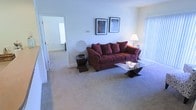 This single-floor residence offers a comfortable and well-appointed living environment designed with everyday functionality in mind. The living room serves as the heart of the space, centered around a plush burgundy sofa accented with coordinating throw pillows. A glass-top coffee table with a sculptural base anchors the seating area, while matching side tables add a cohesive, finished look. Two framed art pieces hang symmetrically above the sofa, contributing a sense of curated style to the neutral wall tones. Soft, wall-to-wall carpeting extends throughout, adding warmth underfoot and a sense of comfort to the overall atmosphere. A floor lamp provides warm ambient lighting, while vertical blinds on a nearby window allow for adjustable natural light and privacy. A decorative medallion accent piece adds an artistic touch to the room's corner. An open doorway leads to an adjacent room, suggesting a naturally flowing and connected floor plan. The overall ambiance is calm, neutral, and inviting — well-suited for both relaxed living and easy personalization.