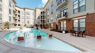 Located at 211 South Greenwood Avenue in Tulsa, Oklahoma, this outdoor amenity area is set within a multi-story residential building distinguished by its striking combination of red brick and neutral stucco facades, large windows, and private balconies. The centerpiece is a spacious resort-style swimming pool with a gradual entry, surrounded by a broad concrete deck furnished with red and white lounge chairs, providing a relaxing retreat for residents. Adjacent to the pool, a generous patio area features wicker sectional seating with vibrant green cushions, offering a comfortable outdoor living space. A covered lounge corridor with exposed brick walls, recessed lighting, and a double-height ceiling connects the interior to the outdoors, creating a sheltered social zone. A dedicated outdoor kitchen area includes a built-in bar counter with bar stools and overhead umbrella seating, ideal for casual dining and entertaining. Paver walkways and manicured landscaping with ornamental shrubs and globe lighting tie the various outdoor zones together cohesively. The courtyard orientation maximizes sunlight throughout the day while the surrounding building provides a sense of enclosure and privacy, making this a well-appointed and functional outdoor community space.