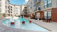 Located at 211 South Greenwood Avenue in Tulsa, Oklahoma, this outdoor amenity area is set within a multi-story residential building distinguished by its striking combination of red brick and neutral stucco facades, large windows, and private balconies. The centerpiece is a spacious resort-style swimming pool with a gradual entry, surrounded by a broad concrete deck furnished with red and white lounge chairs, providing a relaxing retreat for residents. Adjacent to the pool, a generous patio area features wicker sectional seating with vibrant green cushions, offering a comfortable outdoor living space. A covered lounge corridor with exposed brick walls, recessed lighting, and a double-height ceiling connects the interior to the outdoors, creating a sheltered social zone. A dedicated outdoor kitchen area includes a built-in bar counter with bar stools and overhead umbrella seating, ideal for casual dining and entertaining. Paver walkways and manicured landscaping with ornamental shrubs and globe lighting tie the various outdoor zones together cohesively. The courtyard orientation maximizes sunlight throughout the day while the surrounding building provides a sense of enclosure and privacy, making this a well-appointed and functional outdoor community space.