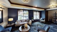 This thoughtfully designed single-floor space offers a refined blend of comfort and sophistication across several distinct areas. The main lounge is anchored by large street-facing windows that flood the room with natural light, offering views of the surrounding neighborhood. Deep navy blue accent walls contrast elegantly with crisp white crown molding and rich dark wood trim, creating a bold yet inviting atmosphere. Plush tufted sofas, accent armchairs, and layered textiles are arranged around round wood coffee tables, lending the space a curated, residential warmth. Geometric patterned carpeting adds visual depth underfoot, while a contemporary chandelier provides stylish overhead illumination. A transitional corridor lined with framed black-and-white photography and classic wainscoting connects the main lounge to a private dining or conference area, which features an arched stone accent wall, warm wood flooring, and pendant lighting over a communal table. A separate enclosed lounge room stands out for its dramatic character, boasting floor-to-ceiling dark wood built-in shelving, a wood-paneled ceiling, leather club chairs, and a statement chandelier — evoking the feel of a private gentlemen's library. Together, these spaces combine to create a versatile, upscale environment ideal for socializing, meetings, or quiet retreat.