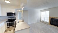 This single-floor, 2-bedroom, 2-bathroom residence offers a well-designed open-concept layout that seamlessly connects the kitchen and living area. The kitchen is equipped with stainless steel appliances, including a range, microwave, and dishwasher, complemented by white cabinetry and light stone-look countertops. A peninsula with a stainless steel sink serves as both a functional workspace and a natural divider between the kitchen and the living space. Track lighting illuminates the kitchen area, while a hallway leads to the bedrooms and bathrooms beyond. The living area features plush carpeting and a neutral color palette, creating a comfortable and inviting atmosphere. A window allows natural light to fill the space, enhancing the bright, airy feel established by the white walls and textured ceilings throughout. The clean, modern finishes and open flow between rooms make this residence a practical and comfortable living environment.