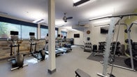 This fitness center is divided into two connected workout zones, accessible through a set of blue double doors with glass panels. The entry area features durable speckled flooring, wall-mounted grab rails, and a mounted exercise mat, providing a functional transition into the main facility.

The cardio zone is bright and well-lit, featuring multiple treadmills, elliptical trainers, and a stationary bike positioned along a textured stone accent wall. Windows fitted with navy blue valances allow natural light to filter in, offering views of the surrounding exterior. Ceiling fans are installed throughout to maintain airflow, supplemented by overhead fluorescent lighting. Wall-mounted flat-screen televisions provide entertainment during workouts.

The strength training zone features a cable machine, weight rack, barbells, and a flat bench, set against a distinctive green accent wall. A full dumbbell rack and a kettlebell station are neatly organized in the free weights area. Motivational framed artwork adds a finishing touch to the space.

The durable, speckled gray flooring runs throughout both zones, with rubber mats placed under equipment for safety. The overall layout is open and functional, offering a comprehensive fitness environment suited to a variety of training needs.