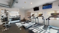 Located in Beverly, Massachusetts, this fitness center offers a comprehensive workout facility designed to support a wide range of exercise routines. The space is laid out across a single floor with a dark rubber flooring throughout, providing both durability and traction during workouts.

The cardio section is well-appointed with multiple treadmills, elliptical trainers, and stationary bikes, arranged to take advantage of the natural light streaming in through several windows. Wall-mounted flat-screen televisions are positioned for easy viewing during cardio sessions. Exposed industrial-style ductwork runs along the ceiling, adding a functional aesthetic to the space.

The strength training area includes a multi-station cable and weight machine, a dedicated free weights section with a full rack of dumbbells in graduated sizes, and additional benches for varied exercises. Workout reference charts are displayed on the walls, offering guidance for users at all fitness levels.

A sliding glass door opens to an outdoor deck area, bringing in additional natural light and views of surrounding greenery. Recessed lighting supplements the natural light throughout. The clean, white walls and open floor plan create an airy, motivating environment well-suited for community fitness use.