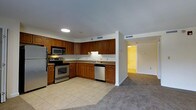 This single-floor space showcases an open-concept layout that seamlessly connects the kitchen and living area. The kitchen is well-appointed with warm medium-toned wood cabinetry providing ample storage, complemented by stainless steel appliances including a refrigerator, range, built-in microwave, and dishwasher. The countertops feature a neutral stone-look finish, and the space is finished with light ceramic tile flooring that adds a clean, practical touch. Recessed lighting illuminates the kitchen area effectively. The adjacent living space transitions to plush carpeting, creating a comfortable and distinct zone for relaxation. A hallway visible in the background suggests additional rooms beyond, with consistent neutral-toned carpeting carrying throughout. The overall color palette is understated and neutral, with the rich wood cabinetry serving as the primary design accent. The layout is functional and efficient, with the open connection between kitchen and living areas promoting an airy, cohesive feel throughout the space.