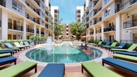 Located at 3115 Routh St in Dallas's vibrant Uptown district, this outdoor courtyard space is the centerpiece of a modern residential complex defined by its striking contemporary architecture. The multi-story building surrounds the courtyard on multiple sides, with clean white facades, black-railed balconies, and large glass doors that frame views of the amenity-rich grounds below.

At the heart of the courtyard sits a generously sized resort-style pool with a decorative fountain feature, surrounded by a spacious deck outfitted with colorful chaise lounges and shaded seating areas. The pool area is framed by mature trees, manicured hedges, and ornamental planters that add greenery and texture to the hardscaped surroundings.

A covered outdoor kitchen and dining area provides a pergola-shaded space equipped with stainless steel grills, a beverage refrigerator, and casual seating beneath a slatted wood overhead structure. Mounted televisions enhance the entertainment functionality of this space.

Pedestrian pathways lined with crushed stone landscaping and mature trees wind through the courtyard, connecting ground-level units—each with private patios enclosed by wrought-iron fencing and lush boxwood hedging—to the broader amenity areas. The overall ambiance is open, social, and well-appointed, offering a vibrant outdoor living experience.