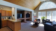This single-floor space makes a strong architectural impression with its dramatic vaulted ceiling clad in warm-toned natural wood planks, which rises to a generous height and immediately draws the eye upward. A prominent full-brick fireplace serves as the focal point of the main living area, flanked by built-in ledges and framed with an arched brick surround. A large flat-screen television is integrated into the brick structure, adding a modern functional element to the classic masonry design.

The living area is carpeted in a neutral patterned textile, providing a comfortable foundation for the seating arrangement, which includes dark upholstered chairs and a wooden coffee table. A large sliding glass door and an arched transom window flood the space with natural light, offering views of an outdoor deck and mature trees beyond.

To one side, a partial kitchen counter with granite-style surfaces and warm wood cabinetry is visible, suggesting an open-concept layout that connects the kitchen to the living space. The overall ambiance is cozy yet spacious, with the interplay of natural wood, brick, and abundant daylight creating an inviting and well-appointed environment.