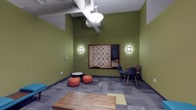 This single-floor recreational lounge is designed as a casual, engaging space with a distinct personality. The walls are painted in a bold olive green, creating a lively and energetic atmosphere throughout the room. A standout feature is the oversized, wall-mounted Scrabble board, framed in dark wood and flanked by two wall sconces that provide warm, focused lighting. Adjacent to the board is a small chalkboard, adding a functional and playful touch.

The space offers a variety of seating arrangements to encourage informal gathering. A pair of rust-orange bean bag chairs and a small round table occupy one corner, while a set of modern navy blue chairs with wooden legs are arranged nearby, providing more structured seating options. A rustic wooden coffee table anchors the center of the room, adding a warm, natural contrast to the space's contemporary elements.

The flooring consists of patterned carpet tiles in varying shades of gray and green, contributing visual interest underfoot. Overhead, exposed ductwork and a pendant light fixture reinforce the industrial aesthetic of the space, balancing the room's playful character with a modern, utilitarian edge.