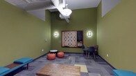 This single-floor recreational lounge is designed as a casual, engaging space with a distinct personality. The walls are painted in a bold olive green, creating a lively and energetic atmosphere throughout the room. A standout feature is the oversized, wall-mounted Scrabble board, framed in dark wood and flanked by two wall sconces that provide warm, focused lighting. Adjacent to the board is a small chalkboard, adding a functional and playful touch.

The space offers a variety of seating arrangements to encourage informal gathering. A pair of rust-orange bean bag chairs and a small round table occupy one corner, while a set of modern navy blue chairs with wooden legs are arranged nearby, providing more structured seating options. A rustic wooden coffee table anchors the center of the room, adding a warm, natural contrast to the space's contemporary elements.

The flooring consists of patterned carpet tiles in varying shades of gray and green, contributing visual interest underfoot. Overhead, exposed ductwork and a pendant light fixture reinforce the industrial aesthetic of the space, balancing the room's playful character with a modern, utilitarian edge.