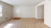 This single-floor unit offers a straightforward and functional layout well-suited for comfortable everyday living. The galley-style kitchen is lined with warm honey-toned wood cabinetry on both upper and lower sections, complemented by light countertops and a double-basin sink. White appliances, including a refrigerator, dishwasher, and range, are neatly integrated into the layout, with a door at the end of the kitchen providing additional access. Light wood-look flooring runs throughout the kitchen, lending a clean, practical surface underfoot.

The living area is open and spacious, featuring neutral carpet and white walls that create a blank canvas adaptable to various furnishing styles. Natural light enters through a window fitted with horizontal blinds, brightening the space. A front entry door opens directly into the living area, which connects seamlessly to the rest of the unit.

The bedroom is a well-proportioned room with the same neutral carpet and white walls, featuring a window that allows natural light to filter in. Multiple electrical outlets are positioned throughout.

The bathroom is clean and utilitarian, featuring a full-size soaking tub with a surrounding white shower enclosure equipped with built-in shelving, alongside a toilet and mirror.