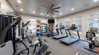 Located at 7805 S Union Ave in Tulsa, Oklahoma, this fitness center offers a comprehensive, single-floor exercise space designed to support a wide range of workout routines. The room is bright and inviting, with multiple windows along the walls that allow abundant natural light to fill the space, complemented by recessed ceiling lighting and a large ceiling fan that promotes air circulation during workouts.

The layout is thoughtfully organized to accommodate both cardio and strength training. A row of treadmills and an elliptical machine lines one side of the room, providing dedicated space for cardiovascular exercise. On the opposite side, a multi-station cable and weight machine offers versatile strength training options. A full rack of dumbbells is neatly arranged for free weight exercises, alongside a TAC-branded rack stocked with kettlebells, medicine balls, and stability balls of varying sizes to support functional fitness training.

Floor-to-ceiling mirrors span one wall, enhancing the sense of space and allowing users to monitor their form. Wall-mounted flat-screen televisions provide entertainment during workouts. Durable rubber flooring covers the exercise area, ensuring safety and comfort. Rustic wooden shelving adds a decorative touch, and a water station is conveniently available. The entrance features a glass-paneled door, maintaining an open and welcoming atmosphere throughout this well-appointed fitness facility.