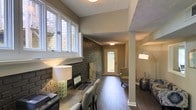 This single-floor space combines character and functionality in a thoughtfully designed layout. A defining architectural feature is the striking exposed dark brick wall that runs along one side, adding texture and an industrial charm that contrasts beautifully with the warm, neutral-toned walls throughout. Rich hardwood flooring flows continuously across the space, lending warmth and cohesion to the overall aesthetic.

The workspace area is positioned near a bank of wide casement windows that allow ample natural light to filter in, creating a bright and productive environment. A built-in or desk-style workstation sits comfortably against the brick wall, accompanied by seating that makes the area both practical and inviting.

Toward the rear, a corridor leads to a glass-paneled entry door that brings in additional natural light and provides a view to the outdoors, enhancing the sense of openness. A cozy seating nook with an upholstered chair is tucked near the entrance, offering a welcoming transition point into the space.

A structural column adds an architectural detail while subtly defining zones within the open layout. The textured ceiling and arc floor lamp further contribute to the space's layered, well-appointed ambiance. Overall, the space balances a professional work environment with a warm and comfortable atmosphere.