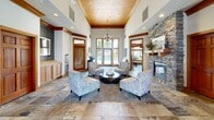Located at 5015 S Regal St in Spokane, WA, this community clubhouse serves as a well-appointed amenity center designed for both relaxation and recreation. The single-floor layout encompasses two living room areas and a dedicated exercise room.

The main living area makes a striking first impression with soaring ceilings, warm wood crown molding, and elegant stone-tile flooring. A floor-to-ceiling stone fireplace anchors the room, complemented by a decorative coffered ceiling with recessed lighting and a chandelier. Large windows and glass doors frame views of the outdoor pool area, flooding the space with natural light. Comfortable seating arrangements invite gathering and relaxation.

The entry foyer continues the refined aesthetic with matching stone tile flooring, tall windows, rich wood-trimmed doorways, and a chandelier overhead, creating a welcoming transition into the facility.

The exercise room is well-equipped with treadmills, stationary bikes, a multi-station cable machine, a power tower, and a dumbbell rack. Natural light filters through multiple windows, and a wall-mounted TV adds convenience during workouts.

Outdoors, a generously sized swimming pool with a wading entry is surrounded by a broad concrete sun deck lined with lounge chairs, shaded seating areas, and lush mature trees, offering a resort-like atmosphere under the open Spokane sky.