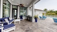 This single-story outdoor amenity space offers a well-appointed covered patio and pool area designed for comfortable leisure and entertaining. The covered patio is finished with large-format stone pavers in a neutral gray tone, creating a polished and durable surface throughout. Tall floor-to-ceiling grid-pane windows and glass French doors line the building's exterior wall, flooding the adjacent interior with natural light while maintaining a clean, modern architectural aesthetic.

The covered lounge area features white-framed outdoor furniture with deep navy cushions, including a full sofa set, accent chairs, and a matching coffee table, all positioned beneath ceiling fans that ensure comfortable airflow. A built-in stone-clad outdoor grill station serves as a functional centerpiece for outdoor cooking and entertaining, complemented by a striking cobalt blue planter with lush tropical foliage.

Beyond the covered area, the space opens to a generous pool deck furnished with blue lounge chairs and side tables arranged poolside. The rectangular swimming pool is framed by a white perimeter fence, with mature palm trees providing a lush tropical backdrop. Additional umbrella-shaded seating is available poolside, completing this resort-inspired outdoor retreat.