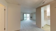 This single-floor residence offers a practical and straightforward layout across several well-defined areas. The main living space is generously proportioned, featuring neutral-toned carpet throughout and a textured ceiling. A sliding glass door leads to a private balcony, allowing natural light to fill the room and providing pleasant outdoor views. A wall-mounted HVAC unit serves the living area, complemented by baseboard heating along the perimeter walls.

A secondary room, likely a bedroom, mirrors the carpeted flooring and features a large window overlooking mature trees, offering a bright and serene atmosphere. A baseboard heater runs along one wall, ensuring consistent climate control.

The kitchen is efficiently designed in a galley-style configuration, outfitted with white cabinetry, granite-patterned countertops, a coil-top electric range, and a full-size refrigerator. Light tile flooring distinguishes the kitchen from the carpeted areas. Track lighting above provides functional illumination. The kitchen connects openly to the living area, which also includes a ceiling fan and access to the unit's entry door.

The bathroom features a full-size soaking tub with a tiled surround and an overhead shower fixture with chrome hardware, offering a clean and functional design.