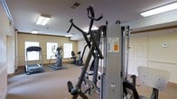 This fitness center is a single-floor facility designed to provide a comprehensive exercise environment for its users. The space is outfitted with a range of workout equipment, including a multi-function cable and strength training machine, an elliptical trainer, a stationary recumbent bike, and a treadmill, offering options for both cardio and resistance training. The equipment is arranged to allow comfortable movement throughout the space, with rubber flooring mats placed beneath machines for safety and stability. The room features neutral-toned walls with classic white trim and paneled doors, giving it a clean and orderly appearance. Recessed fluorescent lighting panels illuminate the space evenly from above, while multiple windows fitted with horizontal blinds allow natural light to filter in, contributing to a bright and energizing atmosphere. The ventilation system is visible through ceiling vents, ensuring proper air circulation during workouts. Overall, the fitness center presents a functional and well-maintained environment suitable for a variety of fitness routines.