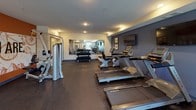 This fitness center is a thoughtfully designed, single-floor facility built to support a comprehensive workout experience. The space is finished with durable rubber flooring throughout, providing both safety and comfort underfoot during exercise. Recessed lighting illuminates the room evenly, creating a bright and energizing atmosphere.

The cardio zone features a row of Matrix treadmills and elliptical machines, ideal for sustained aerobic training. A mounted flat-screen television is positioned along one wall for entertainment during workouts. Adjacent to the cardio area, a dedicated strength training section includes a multi-station cable machine and a rack of dumbbells in graduated weights, catering to a range of fitness levels.

A standout feature is the bold, curved accent wall painted in a rich burnt orange, adorned with a large motivational graphic mural reading "The Stronger You Are, The Better You Feel," giving the space an energetic and inspiring character. A full-length mirror further opens up the room visually and supports proper form during exercise.

A separate functional training area houses medicine balls, resistance accessories, and a yoga mat, offering space for stretching and dynamic workouts. A window along one wall allows natural light to filter in. The overall design balances practicality with visual appeal, making this fitness center a well-rounded destination for daily exercise.