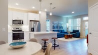 This well-appointed single-floor residence in Davenport, FL offers a thoughtfully designed open-concept layout that seamlessly connects the kitchen, dining, and living areas. The kitchen is outfitted with white shaker cabinetry, light quartz countertops, a subway tile backsplash, stainless steel appliances including a French door refrigerator, electric range, microwave, and dishwasher, along with a central island with bar seating and pendant lighting overhead. Light wood-look flooring runs throughout the home, complementing the neutral color palette and recessed lighting that keep the space bright and airy. The dining area sits near the entry, where natural light filters in through blinds, and a stylish credenza with a round gold mirror adds a decorative focal point. The living room offers generous space with large windows. The bedroom features a bold teal accent wall, ceiling fan, and ample natural light from multiple windows. The en-suite bathroom includes white cabinetry with quartz counters and wood-look flooring. A second full bathroom mirrors this clean aesthetic. A dedicated home office with large windows and a ceiling fan provides a bright, functional workspace. This home combines modern finishes with a coastal-inspired palette throughout.