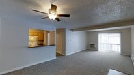 This single-floor space features an open-concept design that seamlessly connects the living area and kitchen. The spacious living room is finished with wall-to-wall carpeting in a neutral tone, creating a comfortable and cohesive feel throughout. A ceiling fan with an integrated light fixture is centrally mounted on the textured popcorn ceiling, providing both airflow and ambient lighting.

The kitchen is partially separated from the living area by a raised pass-through counter, offering a functional division while maintaining an open feel. Warm wood-toned cabinetry lines the kitchen walls, complemented by a white electric range and additional appliances. The counter space provides practical workspace for meal preparation.

A wall-mounted air conditioning unit is positioned near the far wall, ensuring climate control within the living space. A sliding glass door fitted with vertical blinds allows natural light to filter into the room and likely provides access to an outdoor area such as a balcony or patio. Baseboard heating elements are also visible, offering an additional heating option. The overall layout is functional, practical, and well-suited for everyday living.