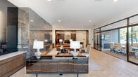 Located at 800 Van Cliburn Way in Fort Worth, Texas, this elegantly appointed lobby spans a generously sized single floor, offering a refined and welcoming first impression. The space is defined by light-toned herringbone hardwood flooring that flows throughout, complemented by soaring ceilings that amplify the sense of openness and grandeur.

At the heart of the lobby, a sophisticated lounge area features curated seating arrangements of leather and textured upholstered chairs, anchored by a large area rug and centered around a striking floor-to-ceiling grey marble fireplace surround that serves as the room's dramatic focal point. A sculptural wood-grain credenza with brass accents and paired table lamps adds warmth and refinement to the reception area.

Transitioning toward the perimeter, a glass-enclosed conference room offers full transparency with floor-to-ceiling glazed walls, flooding the space with natural light and providing panoramic views of the landscaped streetscape outside. The conference room is equipped with a long meeting table, executive seating, and a wall-mounted display screen.

A bold, oversized yellow sculptural chandelier anchors a secondary lounge nook near the building's exterior glass facade, where additional seating and patterned area rugs create an inviting gathering spot. Dark bronzed column cladding and warm wood wall paneling provide striking contrasts against the bright white walls, rounding out this polished, contemporary lobby.