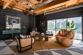 This thoughtfully designed single-floor space masterfully combines industrial and organic aesthetics into a cohesive, inviting environment. The ceiling is a defining architectural feature, showcasing exposed wooden box beams paired with visible structural elements, track lighting, and suspended pendant fixtures that add both function and character. Dark, charred-wood-finish walls create a dramatic contrast against the warm timber overhead, lending the space a bold, contemporary edge.

Large glass garage-style doors span one wall, opening the interior to an outdoor area and flooding the space with natural light and views of lush greenery. A geometric patterned tile floor in varying shades of grey adds visual depth and anchors the open layout. The space is divided into distinct functional zones: a dining area features a long dark table surrounded by eclectic floral-upholstered chairs, while a built-in counter along one wall is accented by tiered living plant walls, adding a fresh, biophilic touch.

A lounge area offers a cozy retreat, complete with a freestanding wood-burning stove, cane-accent armchairs, and eclectic decorative art pieces. A sunlit reading nook near oversized windows features unique woven furniture and tropical potted plants. A projector mounted to the ceiling suggests versatile presentation or entertainment capabilities, making this space equally suited for social gatherings, collaborative work, or relaxed leisure.