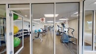The fitness center at Villas At Pine Hills, located at 1 Country Club Drive in Manorville, New York, is a thoughtfully designed single-story amenity facility managed by Heatherwood. Entry is through glass-paneled doors that open into a clean, tile-floored lobby area, which includes a water cooler station and restroom access, providing convenience for visitors before and after workouts.

The main workout floor is expansive and divided into distinct functional zones. The cardio section features a generous row of treadmills and elliptical trainers, all positioned to allow comfortable spacing between users. Large panel lighting overhead ensures consistent, bright illumination throughout. The strength training area is well-stocked with a full dumbbell rack, adjustable weight benches, stability and medicine balls, and a variety of cable and resistance machines. Full-length mirrors line portions of the walls, enhancing the sense of space and allowing users to monitor their form.

Durable speckled rubber flooring covers the workout areas, offering both safety and resilience. Windows along the perimeter allow natural light to filter in and provide views of the surrounding outdoor environment. A wall-mounted television adds to the amenity's functionality. The overall atmosphere is clean, organized, and well-maintained.