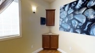This dedicated massage room offers a thoughtfully designed environment tailored for professional therapeutic services. The space features warm, neutral-toned walls that promote a calm and relaxing atmosphere, complemented by light hardwood flooring that adds a natural, inviting quality to the room.

A standout functional element is the corner vanity unit, crafted with rich wood cabinetry and topped with a granite-style countertop housing an integrated sink — an essential and practical feature for a professional treatment space. Above the vanity, a matching upper wall cabinet provides convenient storage for supplies and linens, keeping the room organized and clutter-free. A wall-mounted paper towel dispenser nearby adds to the hygienic functionality of the setup.

Soft, ambient lighting is provided by an elegant wall sconce, casting a warm and gentle glow that enhances the overall soothing ambiance. Decorative wall art featuring smooth, cool-toned stones reinforces the tranquil, wellness-focused theme of the space. The clean, uncluttered design of the room ensures the focus remains on comfort and relaxation, making it well-suited for massage therapy or other personal wellness treatments.