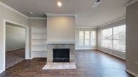 This single-story home offers a thoughtfully designed layout with consistent dark hardwood-style flooring and neutral tones throughout. The spacious living room serves as the heart of the home, anchored by a tile-surround fireplace with a white mantel and flanked by built-in open shelving. Multiple windows allow generous natural light to fill the room, creating a bright and welcoming atmosphere. Crown molding adds a refined architectural touch to the main living areas.

The kitchen is well-equipped with rich, dark shaker-style cabinetry, light granite countertops, a white subway tile backsplash, and a full suite of stainless steel appliances. A row of windows along the kitchen wall provides outdoor views and additional natural light, making it an inviting space for cooking and gathering.

Arched doorways connect the hallways, lending a classic architectural character to the interior. The primary bathroom features a lengthy granite-topped double vanity with dark cabinetry, a large mirror, and direct access to a walk-in closet with wire shelving. A full bathtub with shower is also present in the bathroom. The dedicated laundry room includes side-by-side washer and dryer units. A rear door with a full-length window provides backyard access from a secondary living or dining area, which also includes a ceiling fan.