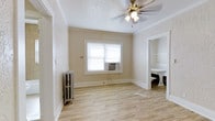 This single-floor space combines vintage architectural character with functional design. The main room features light wood-tone vinyl flooring that runs throughout, creating a warm and cohesive look underfoot. Neutral beige walls with a subtle textured finish lend a classic feel, while crisp white crown molding and baseboards provide clean, elegant framing throughout.

A ceiling fan with integrated lighting is centrally mounted, offering both air circulation and ambient illumination. Natural light enters through a double-hung window fitted with a window-unit air conditioner, ensuring year-round comfort. A cast-iron radiator positioned beneath the window speaks to the building's classic construction and provides supplemental heating.

An adjoining area, visible through a wide doorway, reveals built-in white shelving — a practical and attractive storage feature. A pedestal-style fixture is also visible in this secondary space, suggesting a utility or workspace function.

The textured white ceiling adds a layer of traditional detail that complements the overall character of the space. The open, unfurnished layout allows versatile use, making the space well-suited to a range of configurations and purposes.