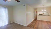 This single-story space presents an open, functional layout that transitions naturally between the living and kitchen areas. The living space features neutral-toned carpet, providing a comfortable and warm underfoot feel, while the adjoining area showcases hardwood-style flooring that extends toward the kitchen. Cream-colored walls throughout create a clean, bright atmosphere, complemented by white baseboards that add a crisp finishing detail.

A ceiling fan is partially visible overhead, offering practical climate control. An overhead HVAC vent is positioned along the upper wall, ensuring efficient air circulation throughout the space. The layout flows openly from the carpeted living area into the hardwood-floored corridor, leading directly to the kitchen, which features wood-toned upper cabinetry and dark countertops. A pass-through or bar-style opening connects the kitchen visually to the adjacent area, enhancing the sense of openness.

The space is straightforward in its design, prioritizing functionality with a neutral palette that offers flexibility for a variety of interior styles and furnishing choices.