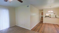 This single-story space presents an open, functional layout that transitions naturally between the living and kitchen areas. The living space features neutral-toned carpet, providing a comfortable and warm underfoot feel, while the adjoining area showcases hardwood-style flooring that extends toward the kitchen. Cream-colored walls throughout create a clean, bright atmosphere, complemented by white baseboards that add a crisp finishing detail.

A ceiling fan is partially visible overhead, offering practical climate control. An overhead HVAC vent is positioned along the upper wall, ensuring efficient air circulation throughout the space. The layout flows openly from the carpeted living area into the hardwood-floored corridor, leading directly to the kitchen, which features wood-toned upper cabinetry and dark countertops. A pass-through or bar-style opening connects the kitchen visually to the adjacent area, enhancing the sense of openness.

The space is straightforward in its design, prioritizing functionality with a neutral palette that offers flexibility for a variety of interior styles and furnishing choices.