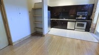 This single-floor space offers an open-concept layout that seamlessly connects the living area to the kitchen. The living space features light hardwood flooring that adds warmth and a clean, bright feel to the interior. A distinctive built-in shelving unit is integrated into the wall alongside a structural column, providing practical open storage while contributing an architectural element to the room.

The kitchen is outfitted with dark espresso-toned cabinetry that contrasts sharply against a black tile backsplash, creating a bold, modern aesthetic. Light granite-style countertops provide a balanced contrast to the darker elements. The kitchen is equipped with stainless steel appliances, including a refrigerator, dishwasher, and range. The flooring transitions from hardwood in the living area to light ceramic tile within the kitchen zone, clearly defining each functional space.

The overall ambiance is modern and functional, with the contrast between dark cabinetry and light flooring creating a visually dynamic interior. The open layout enhances the sense of space, making the unit feel airy and well-connected throughout.